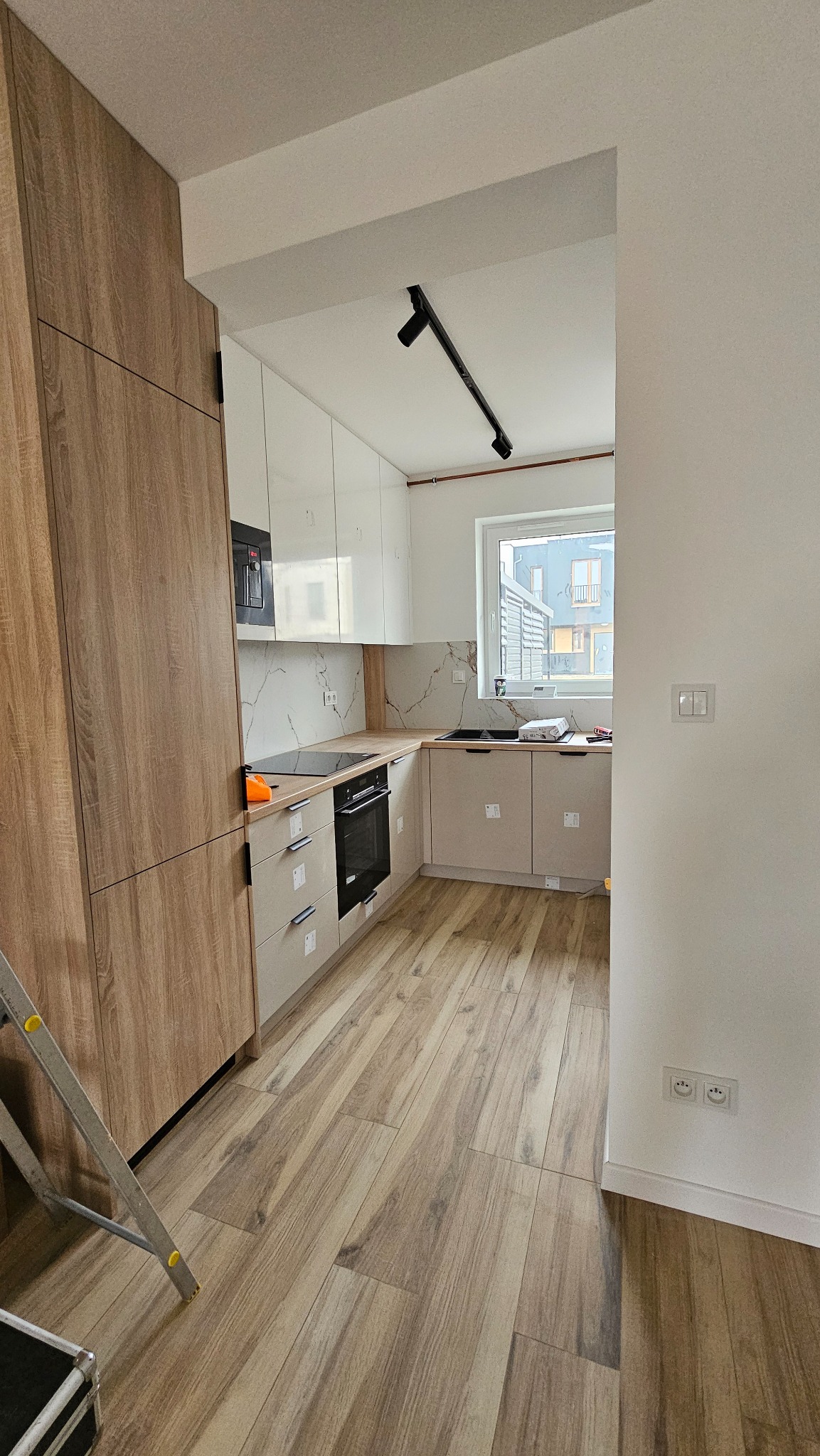 Wnętrze kuchni w trakcie prac wykończeniowych: widoczne nowe panele podłogowe imitujące drewno, zabudowa kuchenna z białymi szafkami górnymi i beżowymi dolnymi, czarny piekarnik i płyta indukcyjna,...