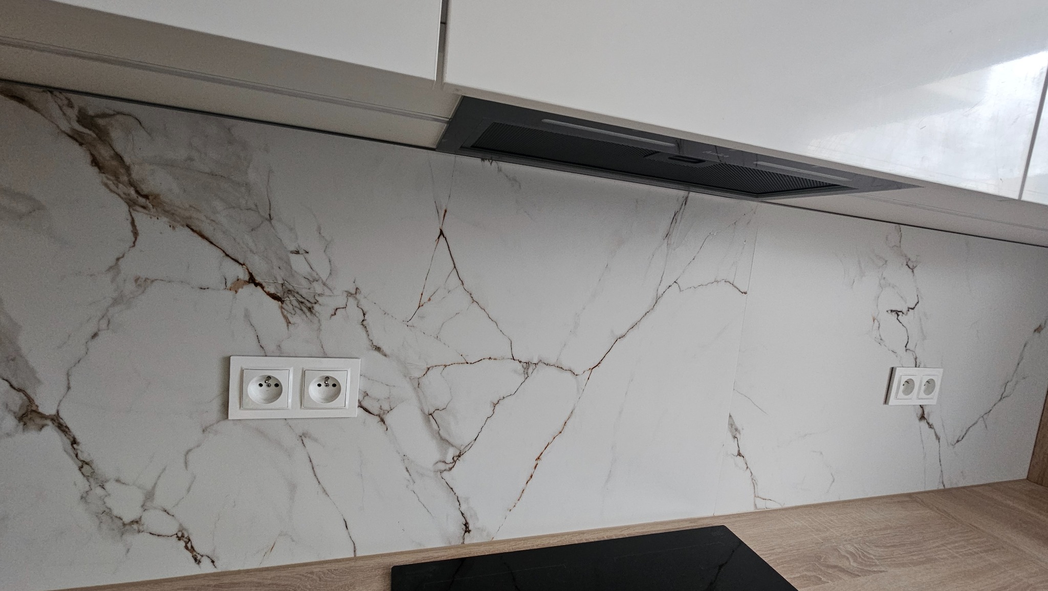 Wykończona ściana nad blatem kuchennym w Zabrzu, imitująca marmur z widocznymi gniazdkami elektrycznymi i fragmentem okapu.
