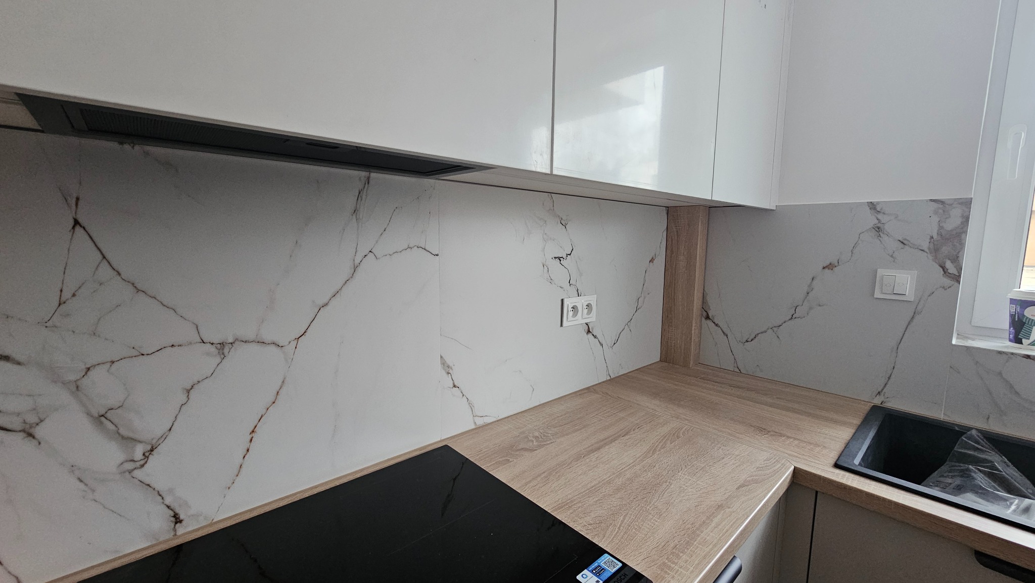 Kuchnia po remoncie: białe szafki wiszące, marmurowe płytki na ścianie, drewniany blat, czarna płyta indukcyjna i zlew.