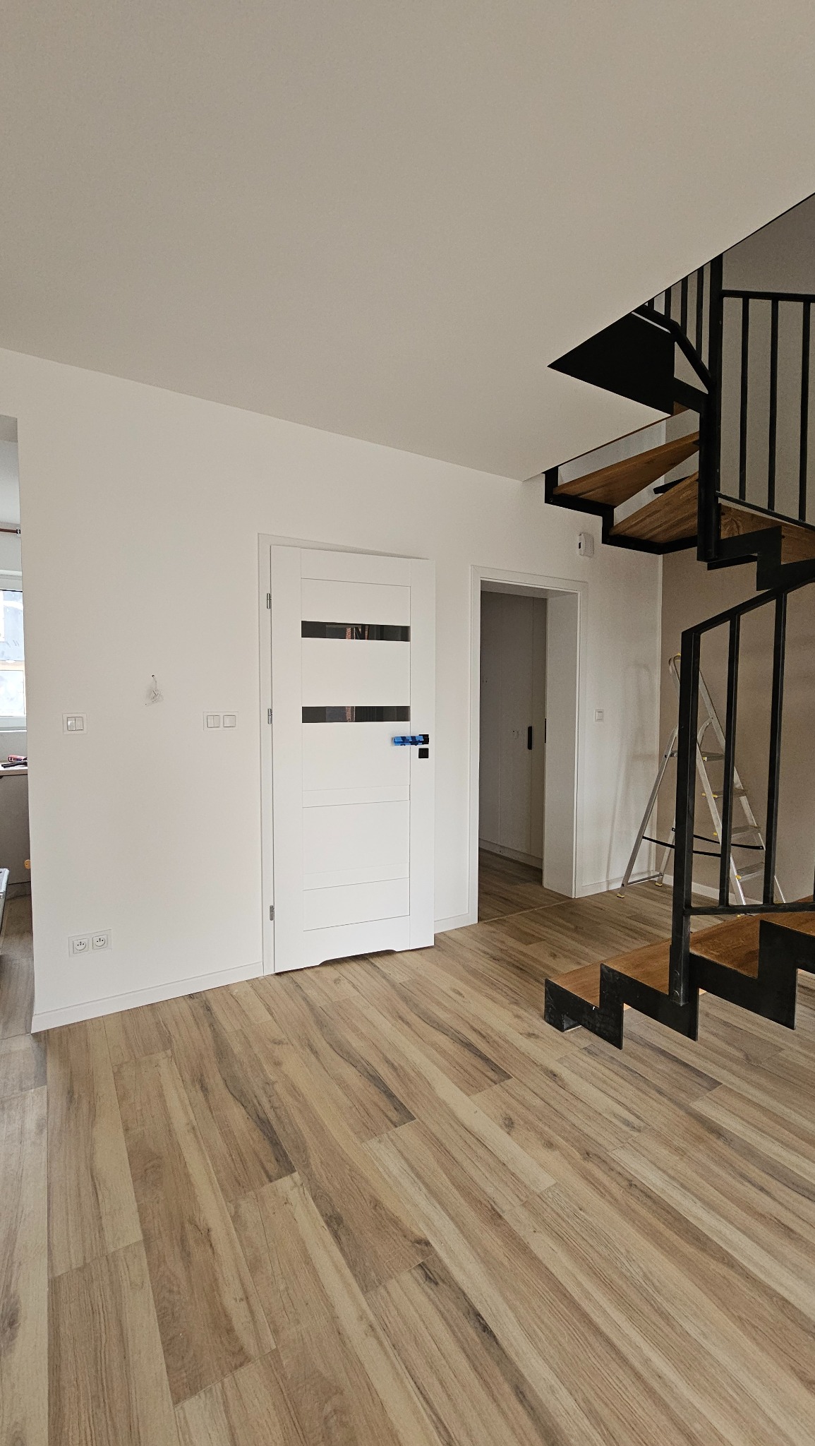 Jasne wnętrze z drewnopodobną podłogą, białą ścianą, nowoczesnymi schodami spiralnymi z metalową konstrukcją i drewnianymi stopniami oraz białe drzwi z poziomymi przeszkleniami, na ścianie widoczne...