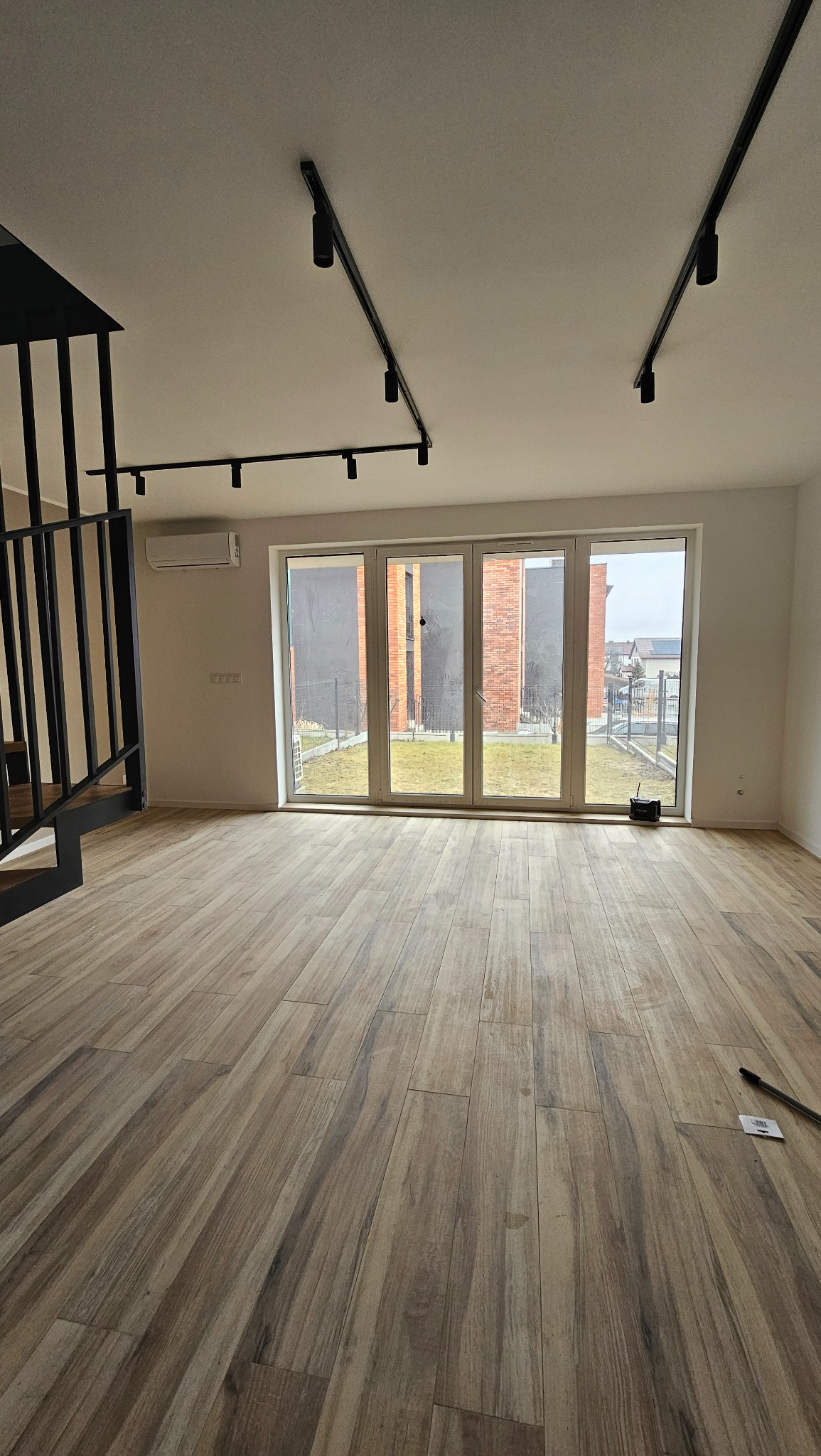 Jasne panele podłogowe imitujące drewno ułożone w pustym pokoju z dużym oknem balkonowym, widoczne czarne szyny oświetleniowe na suficie i czarna metalowa balustrada schodów.