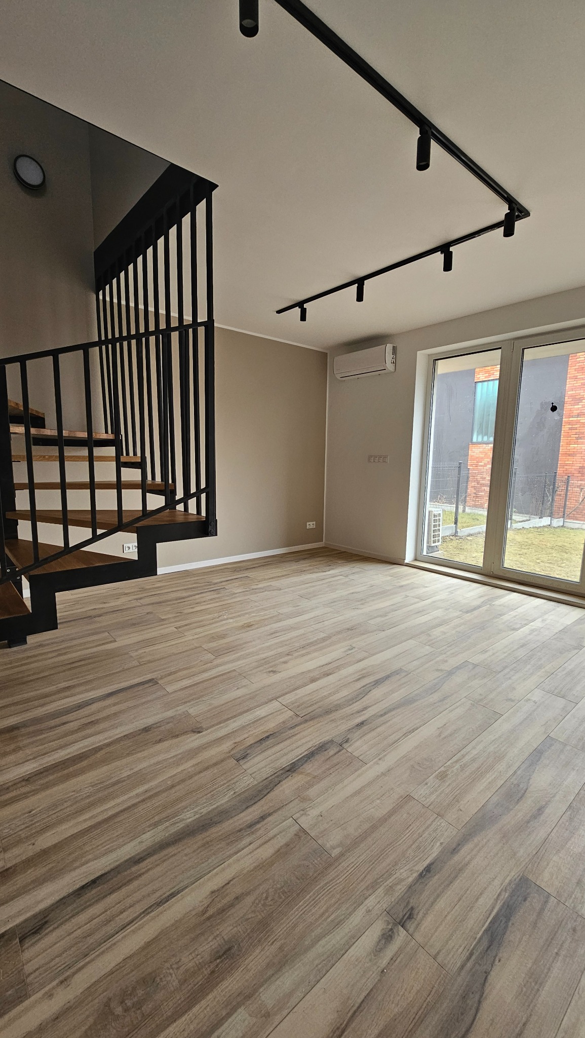 Nowoczesny salon z drewnopodobną podłogą, czarnymi schodami z drewnianymi stopniami, oświetleniem szynowym i klimatyzacją, widok na ogród przez duże okno.