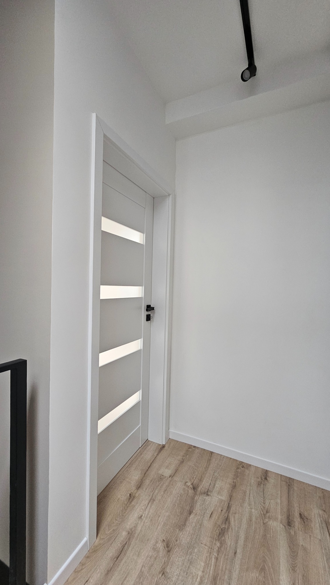 Nowoczesne, białe drzwi wewnętrzne z poziomymi przeszkleniami, czarna klamka, drewnopodobne panele podłogowe, białe ściany i sufit z czarnym reflektorem punktowym.