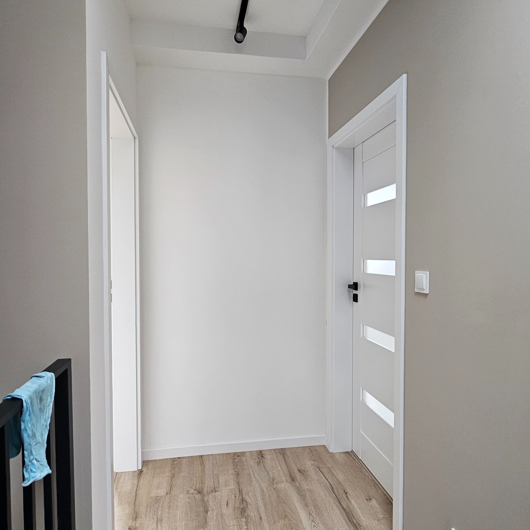 Korytarz po remoncie z drewnopodobną podłogą, białą ścianą, szarą ścianą z włącznikiem, białymi drzwiami z czarną klamką i otwartymi białymi drzwiami z widocznym fragmentem niebieskiej szmatki...