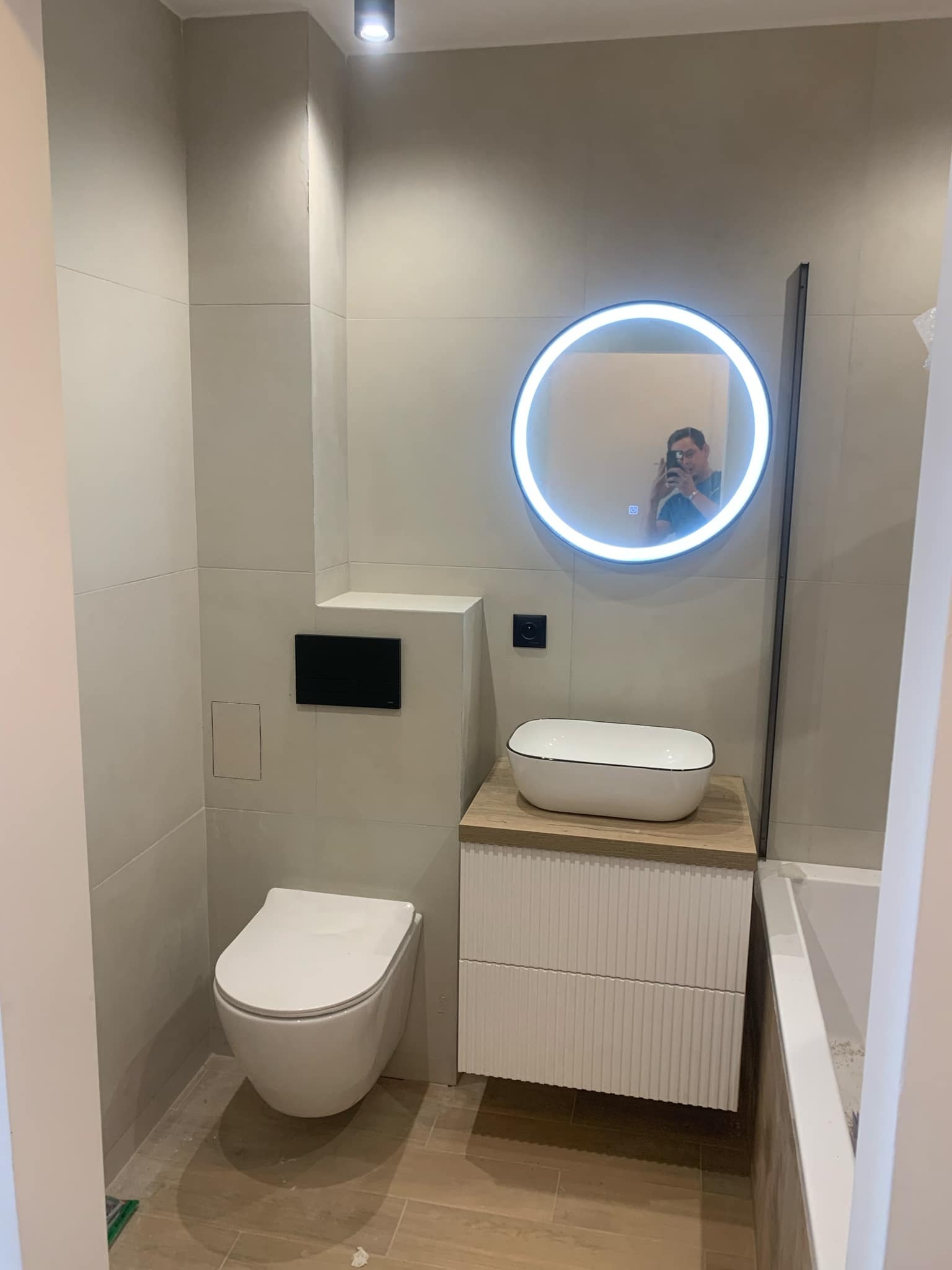 Nowoczesna łazienka z podwieszaną toaletą, umywalką nablatową na szafce z ryflowanymi frontami i okrągłym lustrem z podświetleniem LED, widoczne płytki imitujące drewno na podłodze i szare płytki...