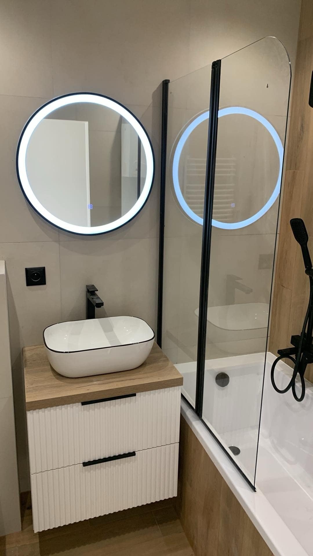 Nowoczesna łazienka z okrągłym lustrem LED, umywalką nablatową na szafce z ryflowanymi frontami i kabiną prysznicową z czarnymi profilami.