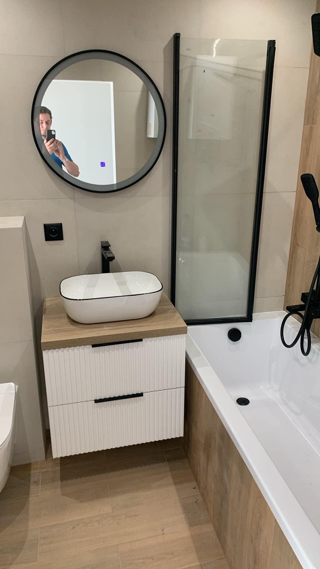 Nowoczesna łazienka z owalną umywalką nablatową z czarnym rantem, baterią w kolorze czarnym, okrągłym lustrem z podświetleniem, kabiną prysznicową z czarnymi profilami i wanną obudowaną płytkami...