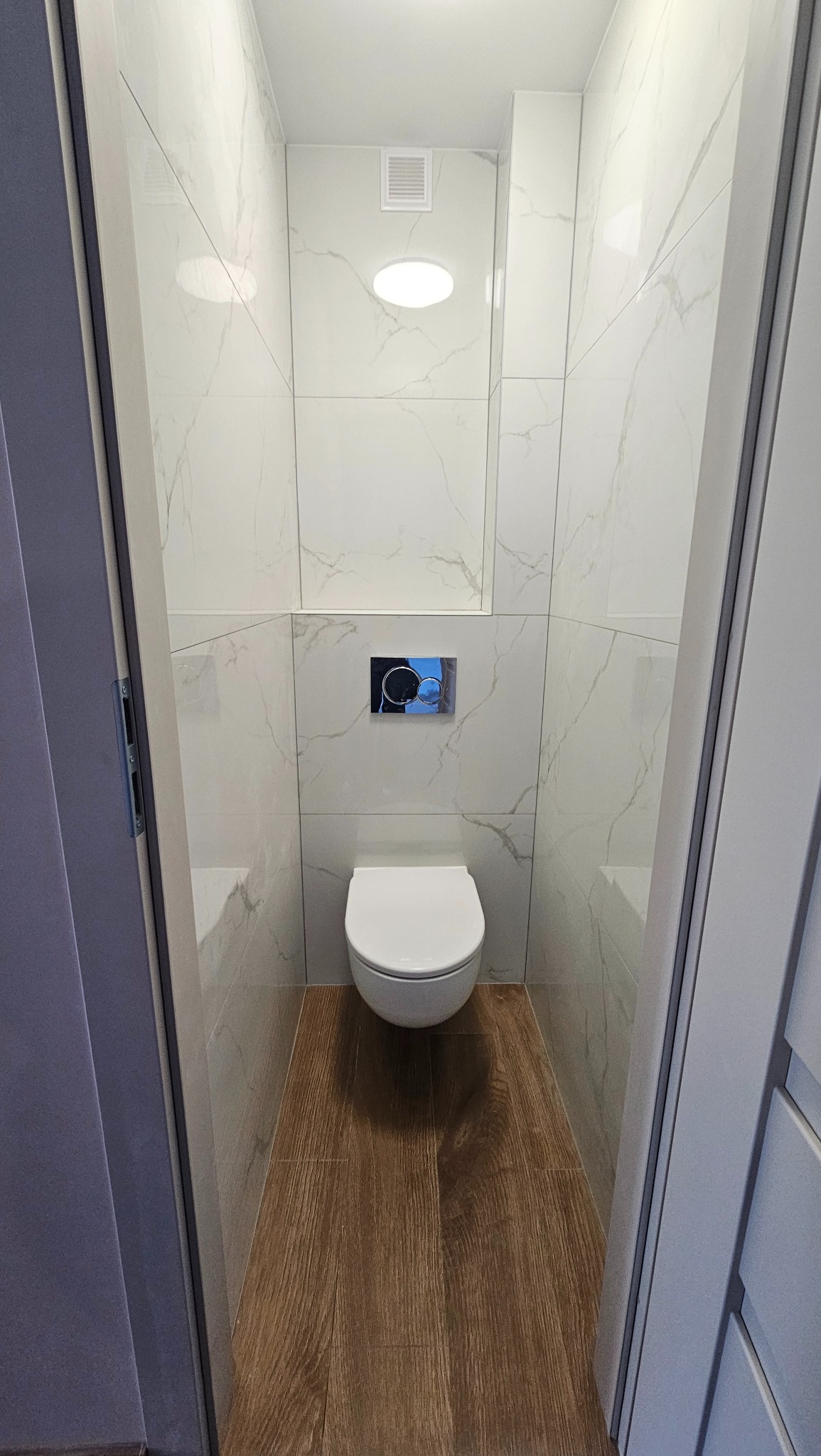 Wąska toaleta po remoncie: białe płytki imitujące marmur na ścianach, drewnopodobne panele na podłodze, podwieszana miska WC i chromowany przycisk spłuczki.