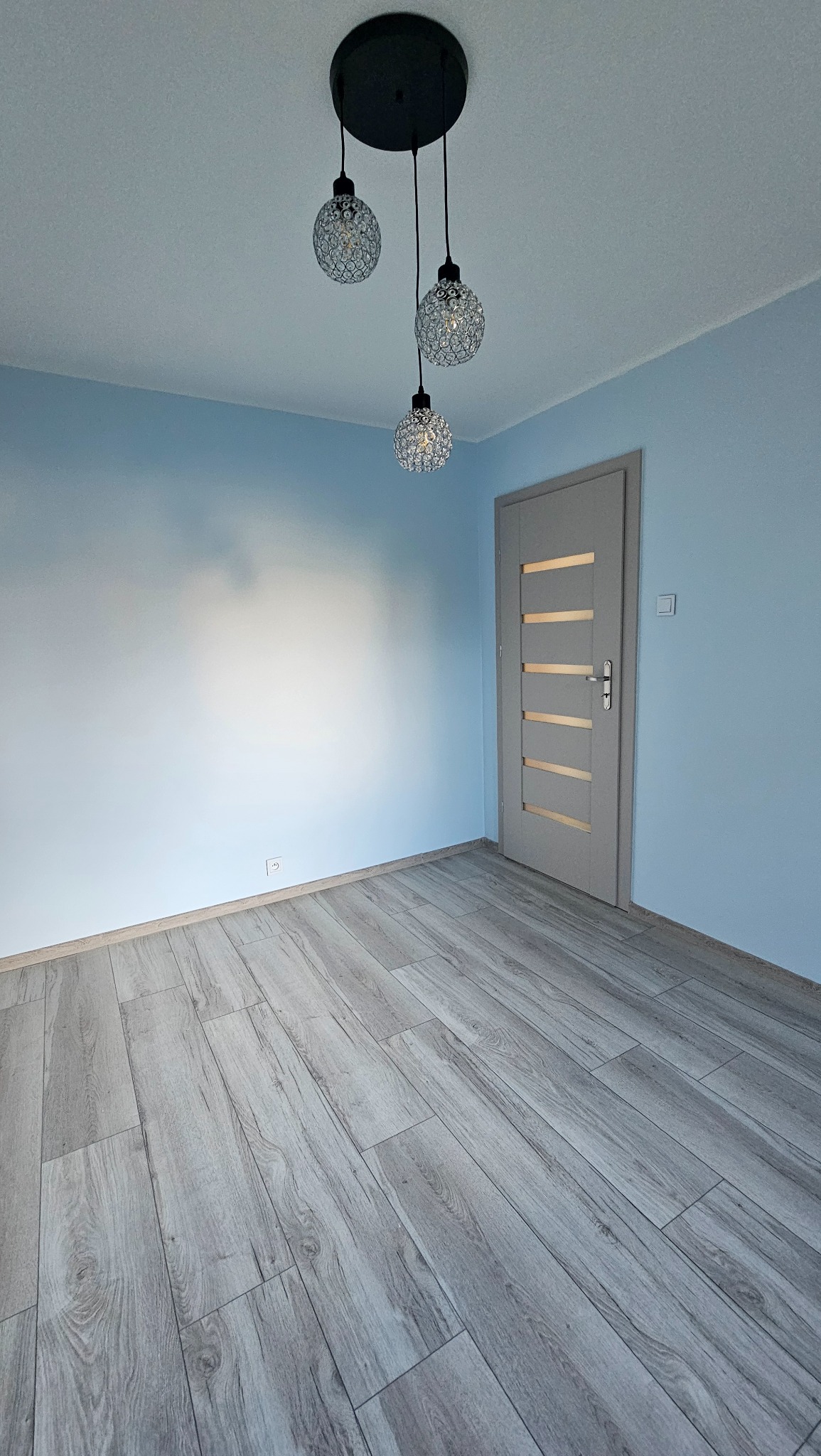 Pomalowane na błękitno wnętrze z szarą podłogą z paneli i szarymi drzwiami z przeszkleniami. Nad podłogą wisi nowoczesna, czarna lampa z trzema kloszami.