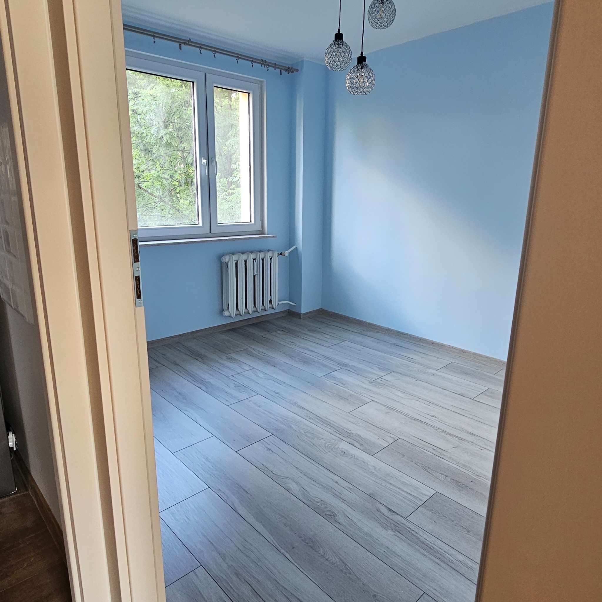 Pomalowany na błękitno pokój po remoncie z panelami podłogowymi w odcieniach szarości, widok zza otwartych drzwi. Na suficie wiszą trzy ozdobne lampy.