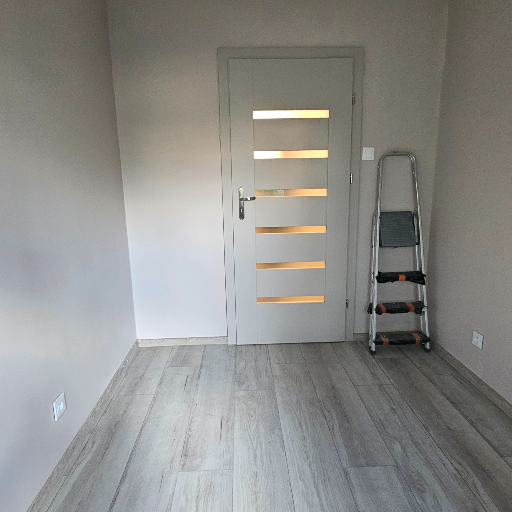 Wnętrze po remoncie: ściany w odcieniach beżu, jasna podłoga z paneli, białe drzwi z przeszkleniami i drabina malarska. Gniazdka elektryczne na ścianach.