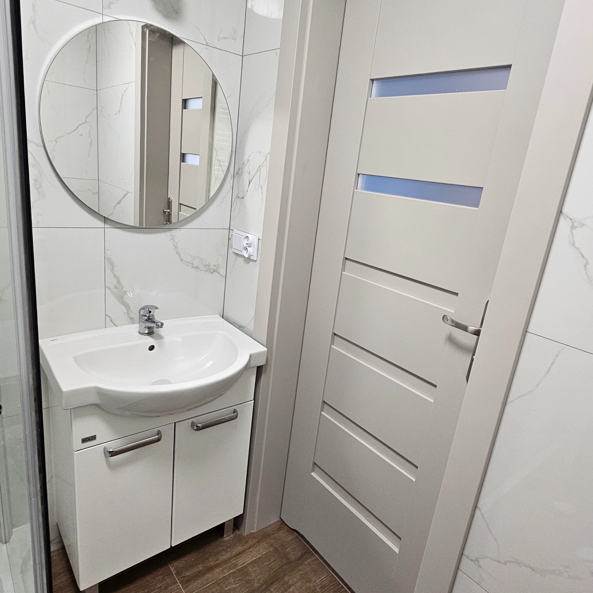 Biała umywalka z szafką Kolo w nowoczesnej łazience z marmurowymi płytkami i drzwiami z poziomymi przeszkleniami. Okrągłe lustro odbija fragment wnętrza.