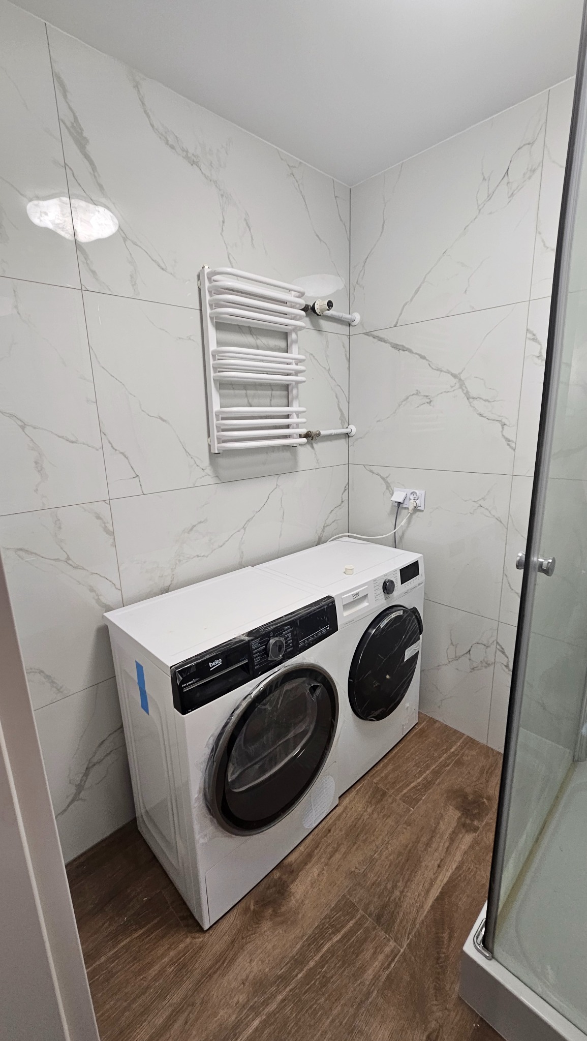Biała pralka i suszarka wkomponowane w wykończoną łazienkę z grzejnikiem drabinkowym w Zabrzu. Płytki imitujące marmur na ścianach, drewnopodobna podłoga.