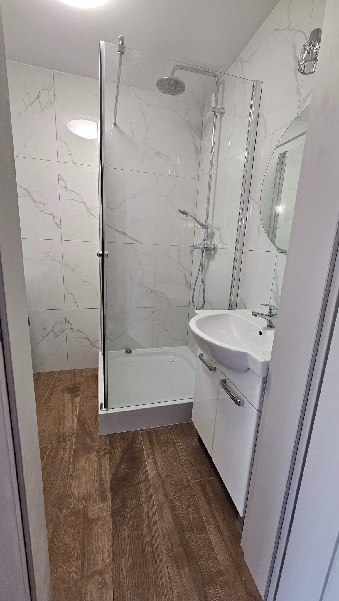 Nowoczesna łazienka po remoncie: kabina prysznicowa ze szklaną ścianką, umywalka z szafką i płytki imitujące marmur na ścianach, drewnopodobna podłoga.