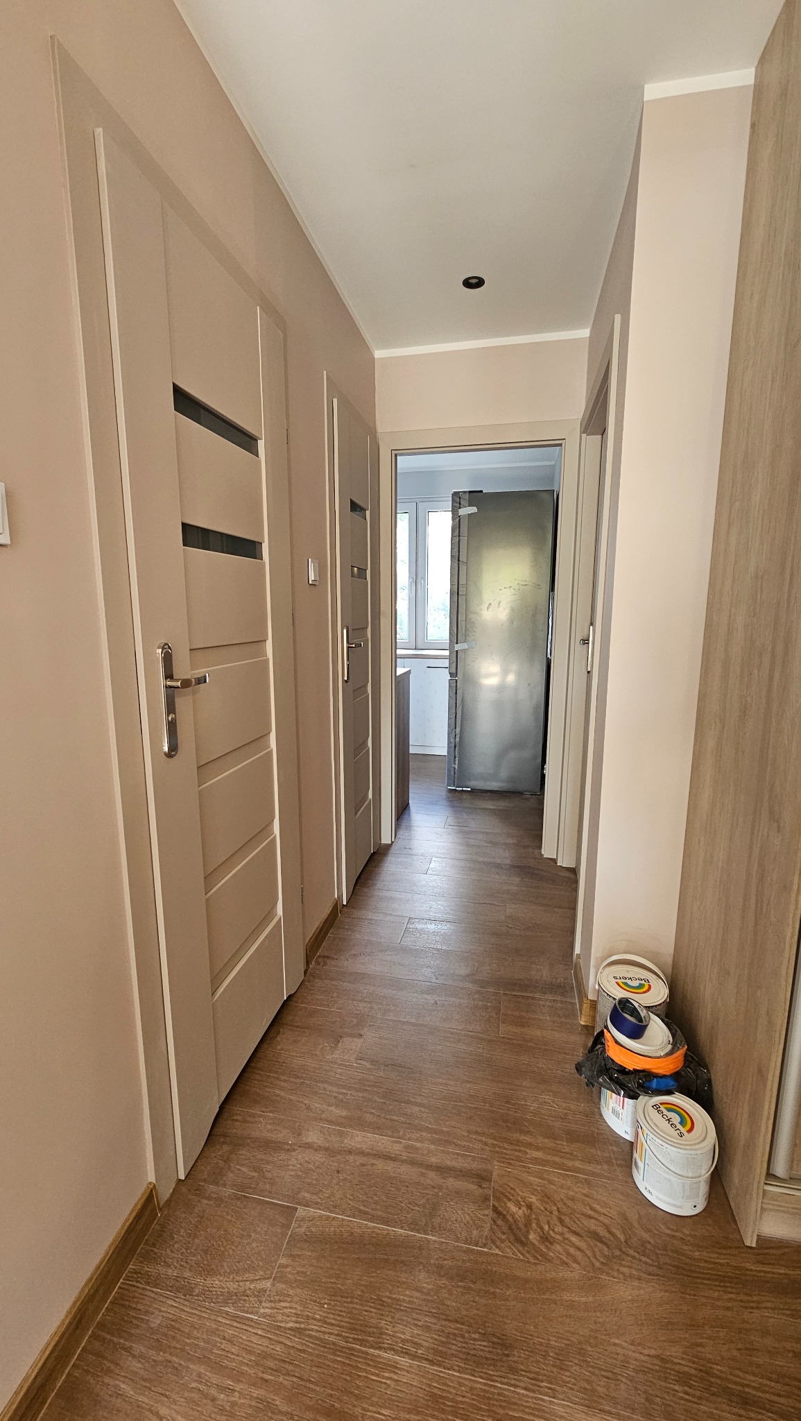 Korytarz po remoncie: drewnopodobne panele, świeżo pomalowane ściany w odcieniach beżu, nowe drzwi z przeszkleniami. Widoczne puszki z farbą.