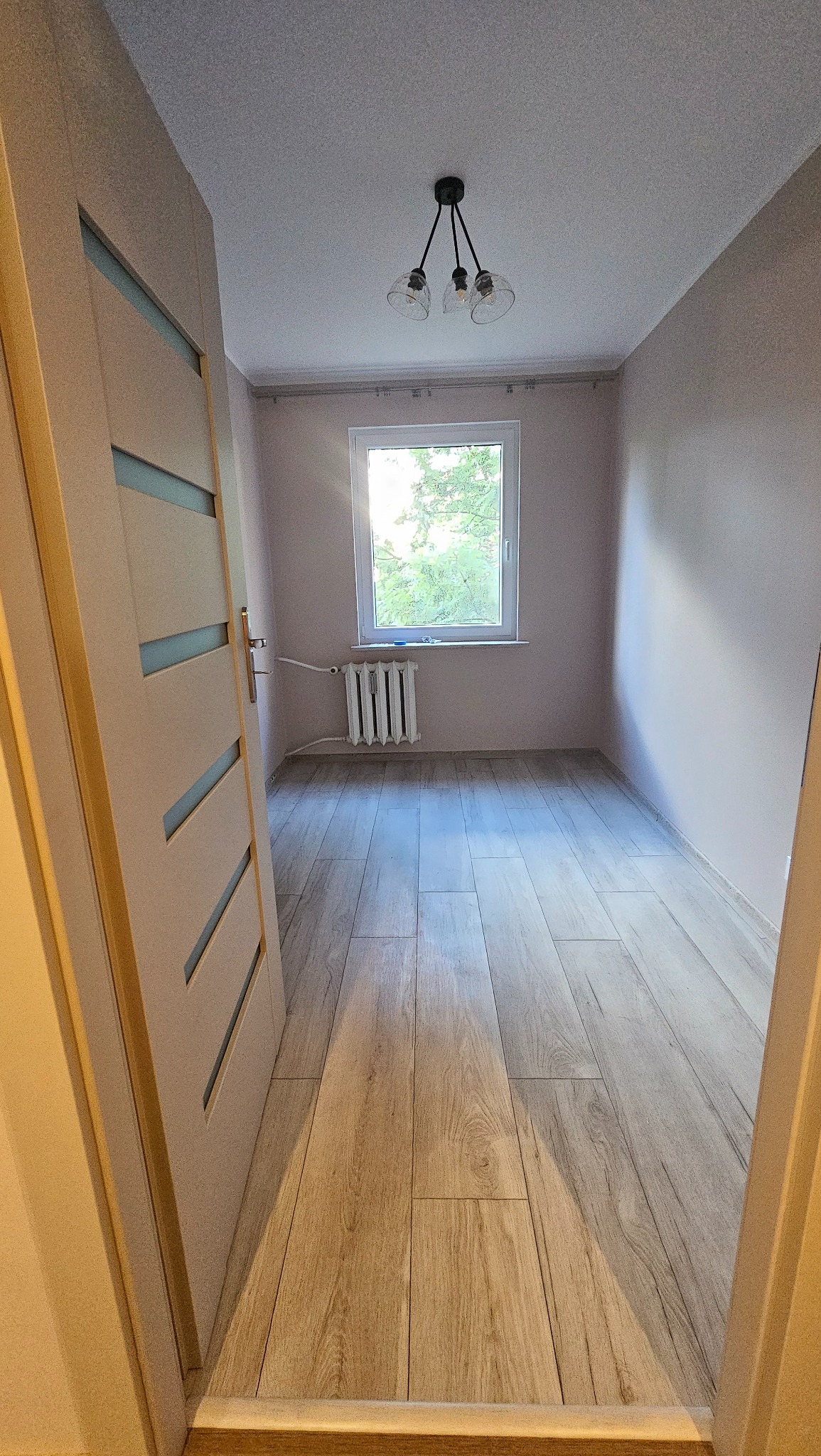 Jasne panele podłogowe imitujące drewno w pokoju z oknem i grzejnikiem. Widok zza otwartych drzwi z przeszkleniami.