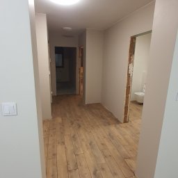 BUD-RIN - Korytarz po remoncie z nową podłogą z paneli drewnopodobnych, widoczne niedokończone wykończenia ścian i futryn, oraz fragment łazienki w głębi.