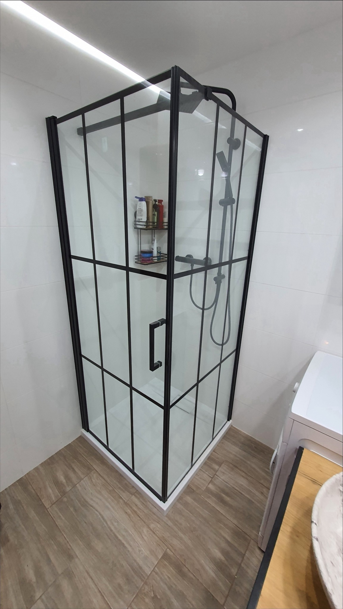 Nowoczesna kabina prysznicowa z czarnymi profilami i szklanymi ściankami, widoczna półka z kosmetykami wewnątrz, jasne płytki na ścianach i podłodze imitującej drewno, oświetlenie LED pod sufitem.