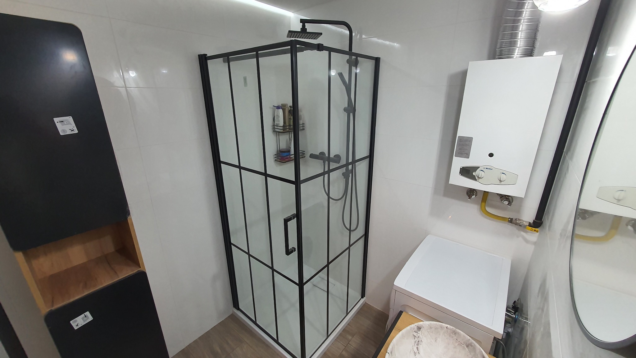 Nowoczesna kabina prysznicowa z czarnymi profilami i deszczownicą w wyremontowanej łazience z białymi płytkami, obok piec gazowy i pralka, a także szafka z otwartymi półkami.