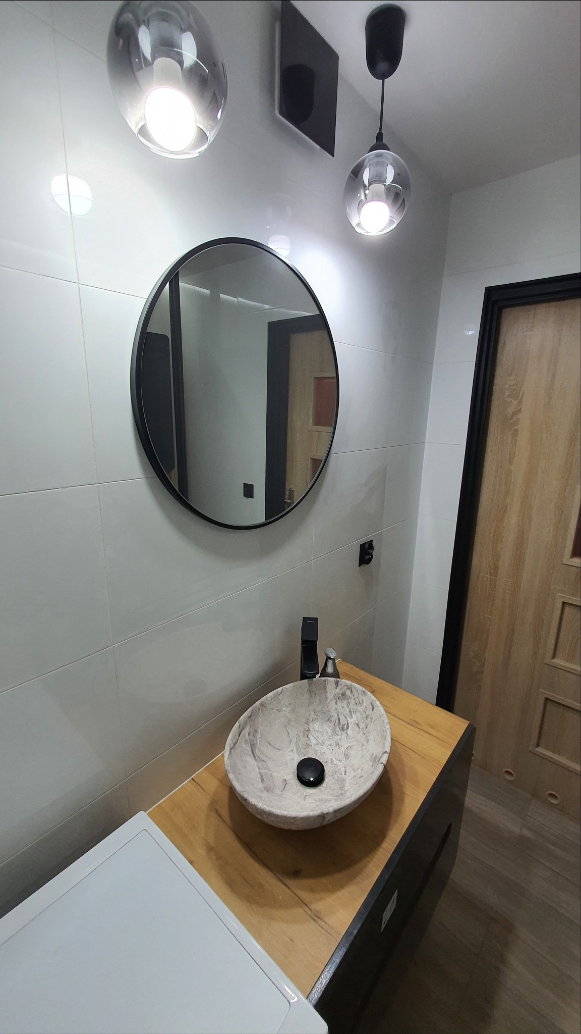 Nowoczesna łazienka z okrągłym lustrem w czarnej ramie, kamienną umywalką na drewnianym blacie, białymi płytkami na ścianach i dwoma szklanymi lampami wiszącymi.
