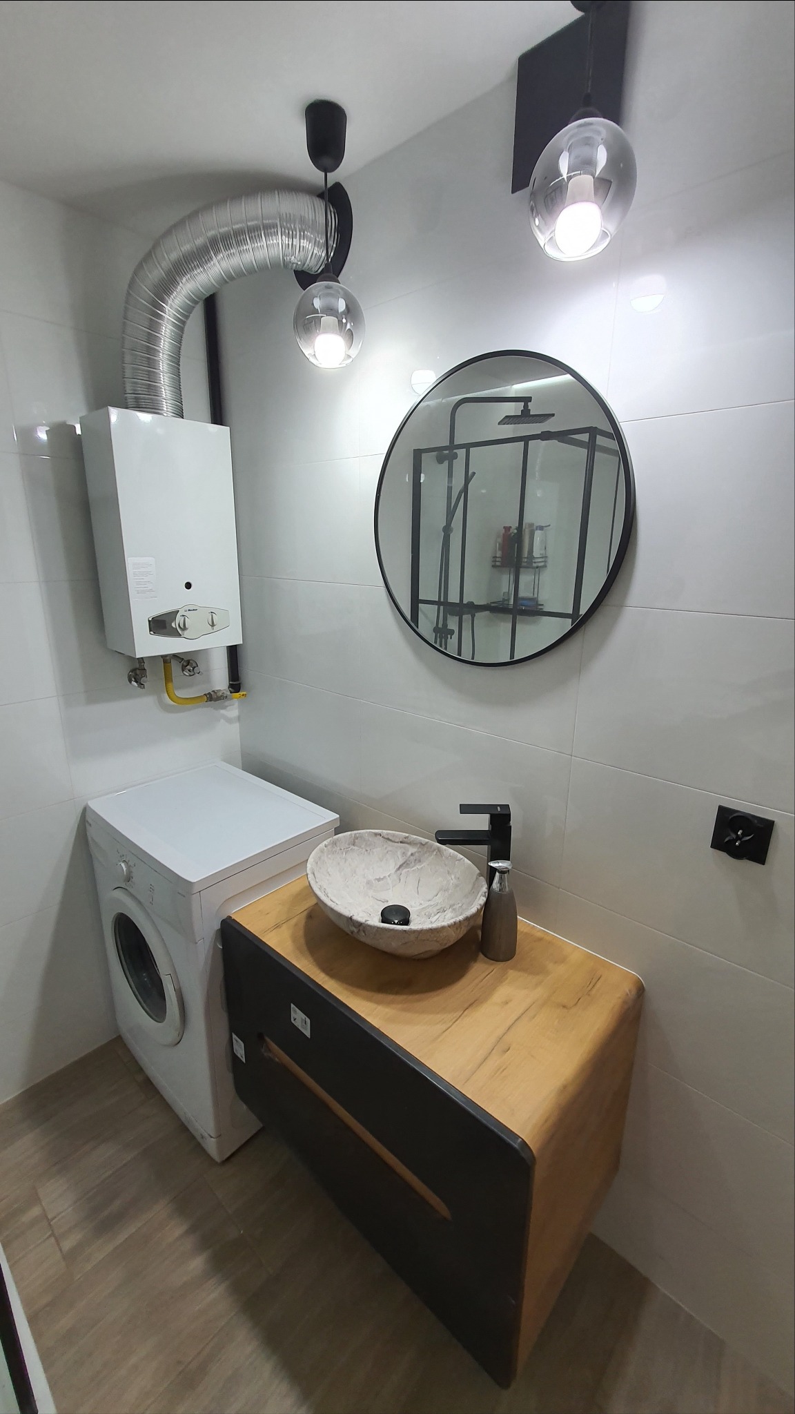 Nowoczesna łazienka z białą armaturą, umywalką przypominającą kamień, okrągłym lustrem z czarną ramą, pralką i piecem gazowym z widoczną rurą odprowadzającą spaliny.