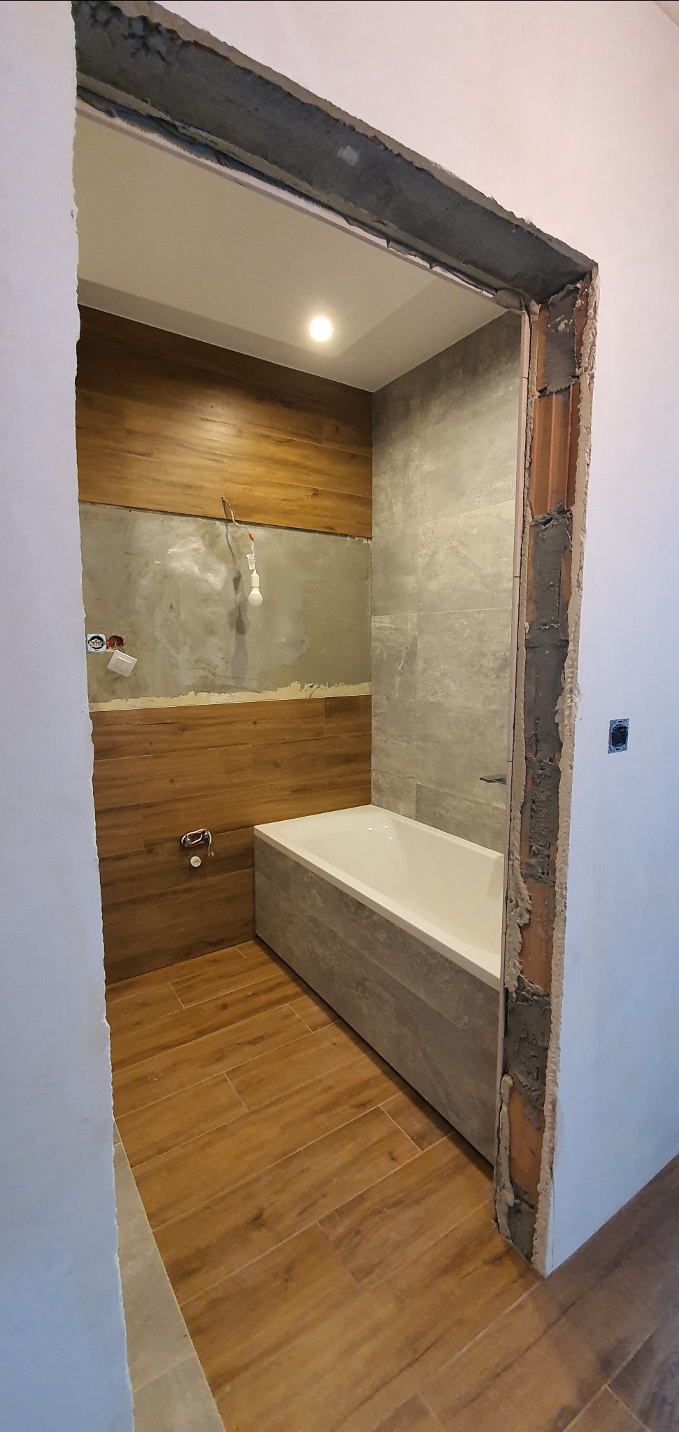 Wnętrze łazienki w trakcie remontu: częściowo wyłożone drewnopodobnymi płytkami ściany, szara płytka na ścianie przy wannie, biała wanna obudowana szarą płytką, niedokończone obramowanie drzwi.