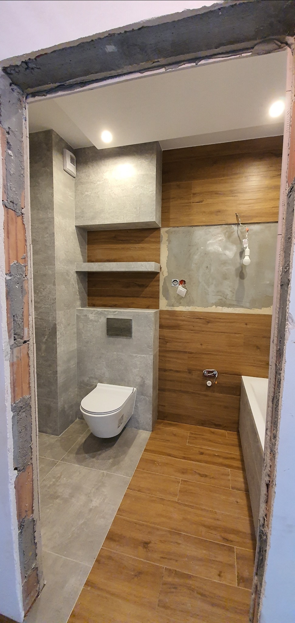 Wnętrze łazienki w trakcie remontu, widoczne płytki imitujące drewno na podłodze i części ścian, szare płytki na pozostałych ścianach, podwieszana toaleta, półki wnękowe, niedokończone przyłącza...