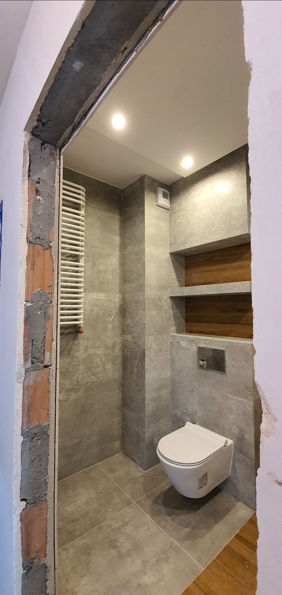 Nowoczesna łazienka w szarościach z wiszącą toaletą i drewnianymi półkami w zabudowie, widok przez surowy otwór drzwiowy z widoczną cegłą i zaprawą.