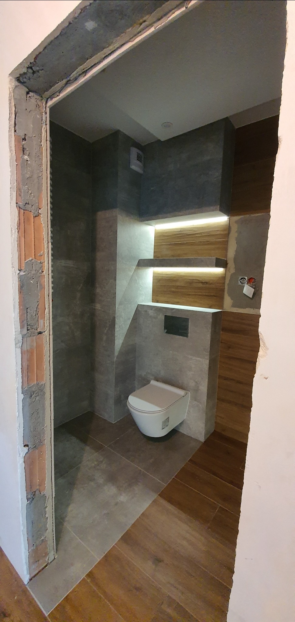 Nowoczesna łazienka w trakcie remontu: podwieszana toaleta na tle szarych płytek, drewnopodobne panele na ścianie, podświetlane półki i surowy mur w ościeżnicy.