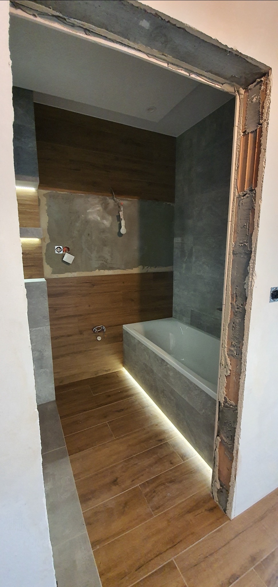 Niedokończona łazienka z drewnopodobnymi płytkami na podłodze i ścianie, podświetleniem LED wzdłuż wanny i podłogi, widoczne instalacje elektryczne i hydrauliczne, surowy framuga drzwi.