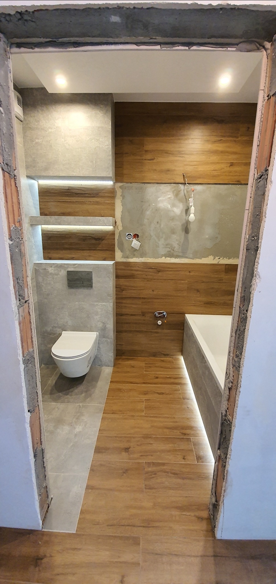 Wnętrze łazienki w trakcie remontu, widok przez surowy otwór drzwiowy, z częściowo położonymi płytkami imitującymi drewno na podłodze i ścianach, oraz szarymi płytkami przy toalecie i wnękach...