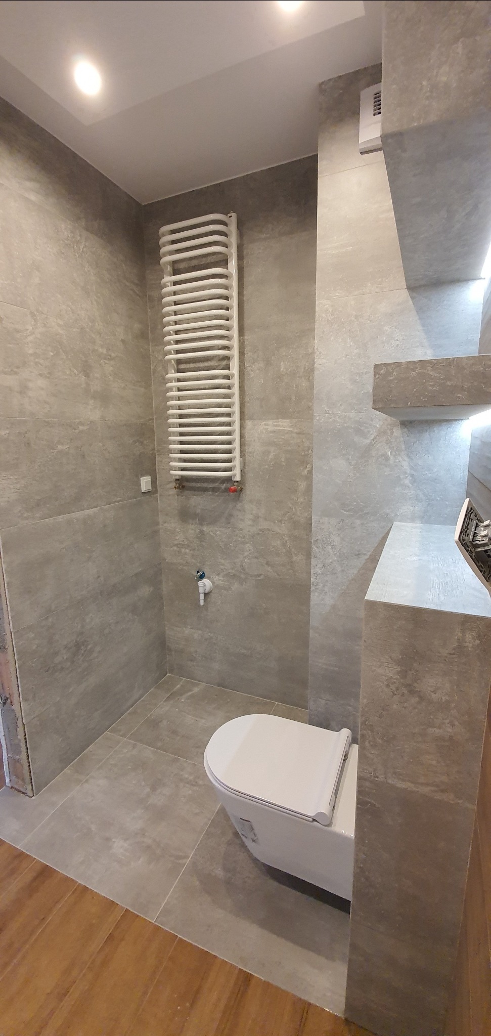 Nowoczesna łazienka w odcieniach szarości z białym grzejnikiem drabinkowym i podwieszaną toaletą, wykończona płytkami imitującymi kamień i z drewnianą podłogą przy wejściu.