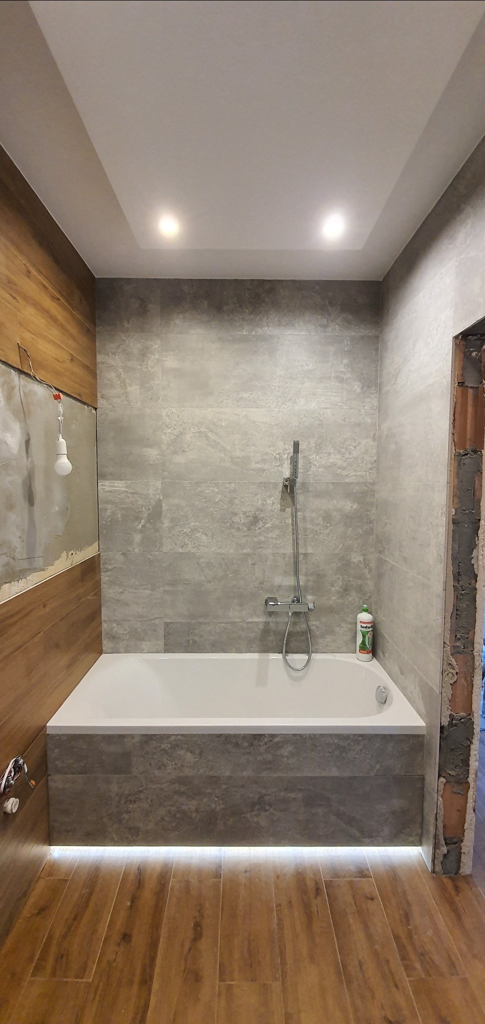 Remontowana łazienka z wanną obudowaną szarą glazurą, drewnopodobną ścianą w trakcie prac wykończeniowych i podświetleniem LED pod wanną.