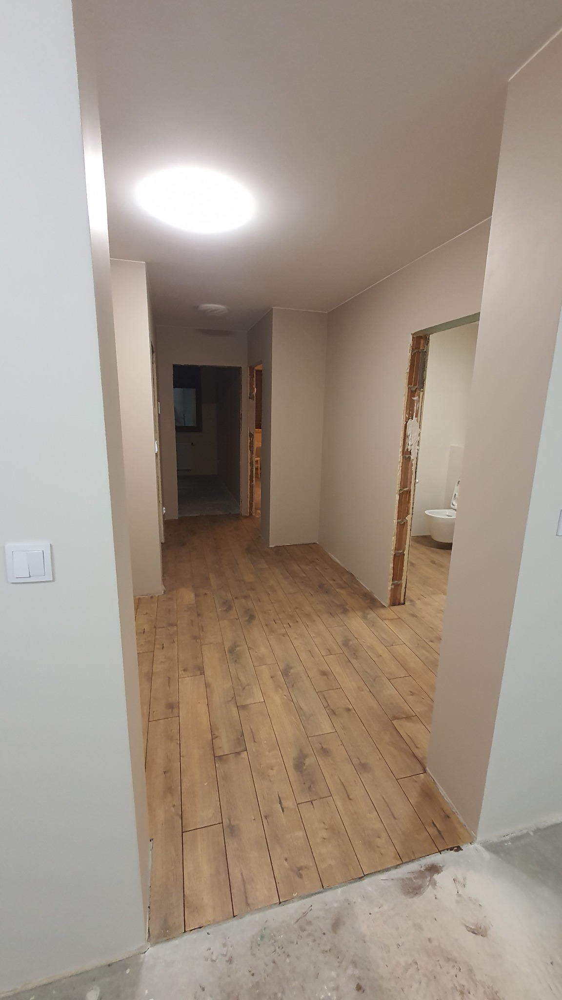 Korytarz po remoncie z nową podłogą z paneli drewnopodobnych, widoczne niedokończone wykończenia ścian i futryn, oraz fragment łazienki w głębi.