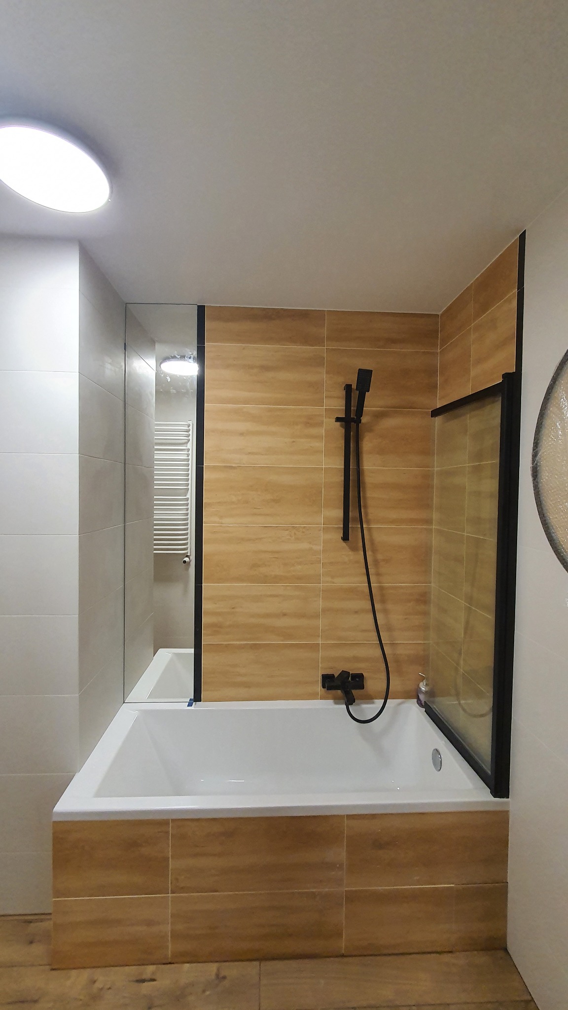 Nowoczesna łazienka z wanną obudowaną płytkami imitującymi drewno, kabina prysznicowa z czarnymi profilami i armaturą w tym samym kolorze.