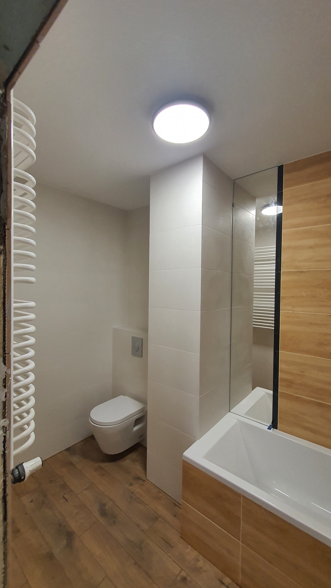 Wykończona łazienka z drewnopodobną podłogą, białymi płytkami na ścianach, podwieszaną toaletą i grzejnikiem drabinkowym w trakcie montażu.