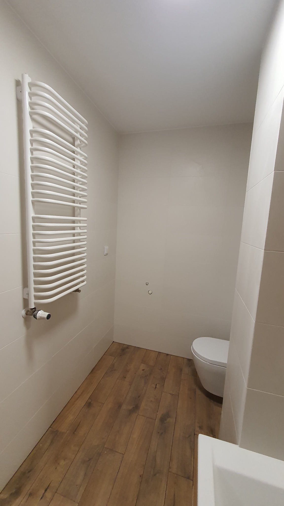 Biała łazienka po remoncie z drewnopodobną podłogą, grzejnikiem drabinkowym i wiszącą toaletą, widoczne ślady po starych mocowaniach na ścianie.