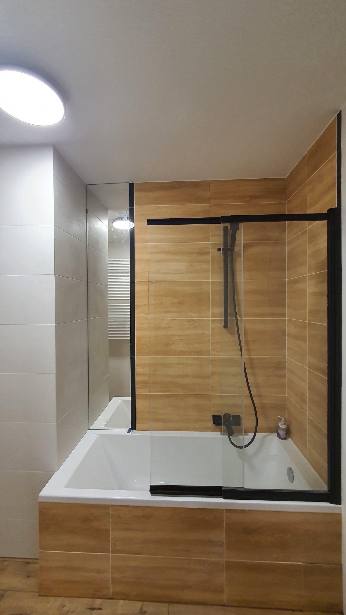 Nowoczesna łazienka z wanną obudowaną płytkami imitującymi drewno, kabina prysznicowa z czarnymi ramami i szklanymi drzwiami przesuwnymi, białe płytki na ścianie obok wanny, widoczny grzejnik...