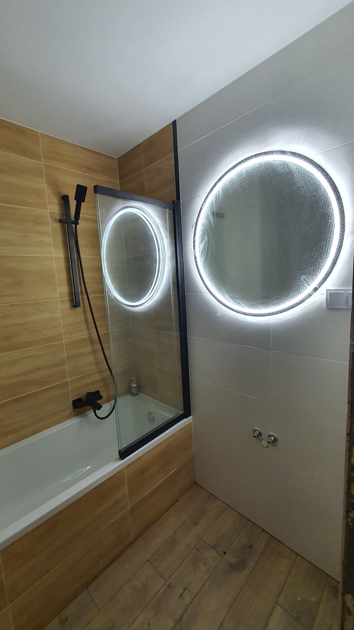 Nowoczesna łazienka z beżowo-białymi płytkami, kabiną prysznicową z czarną ramą i okrągłym lustrem LED. Widoczne drewnopodobne płytki podłogowe i czarny zestaw prysznicowy.