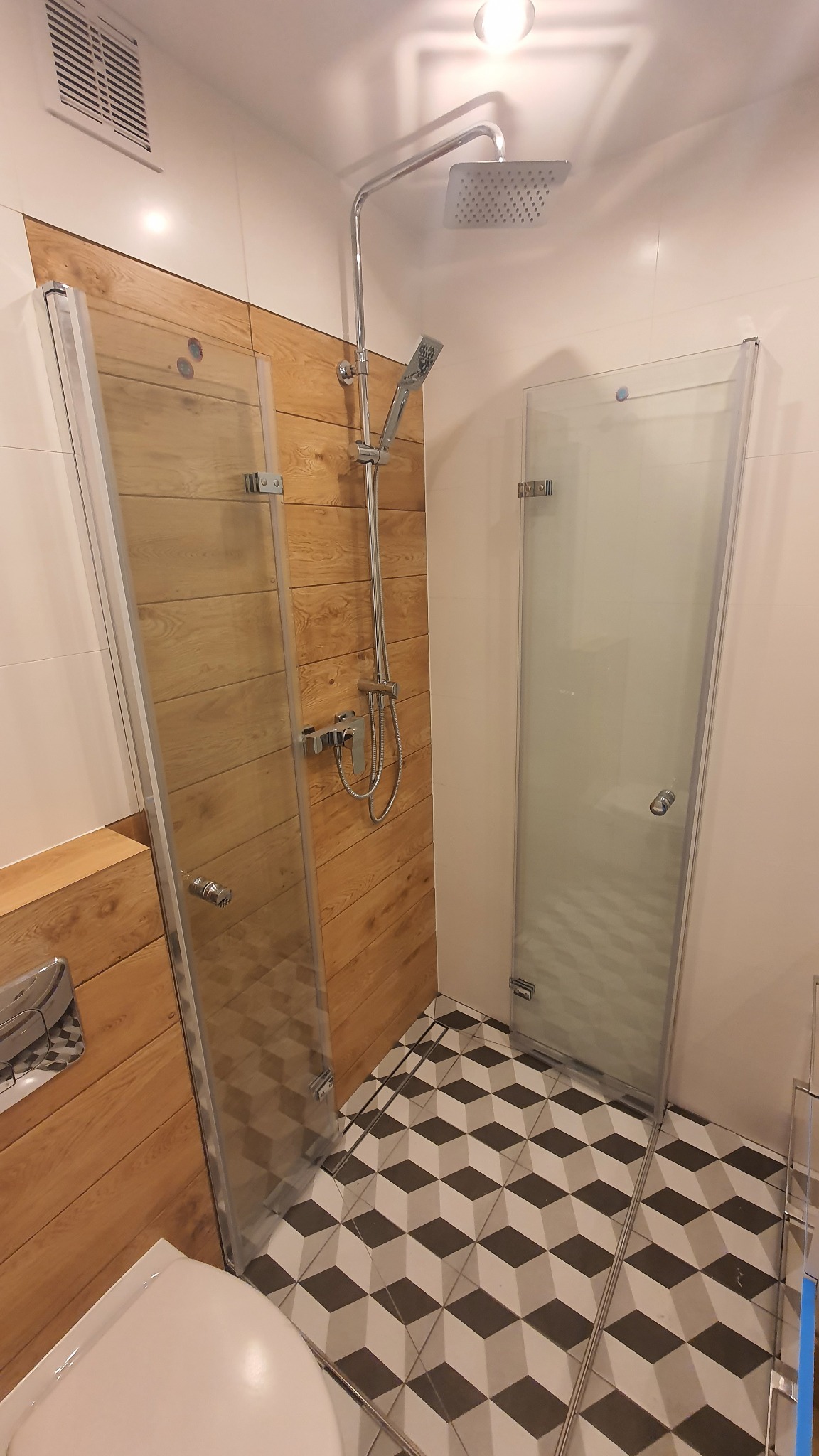 Nowoczesna kabina prysznicowa z dwoma szklanymi ściankami, deszczownicą i panelami imitującymi drewno, na podłodze wzór geometryczny.