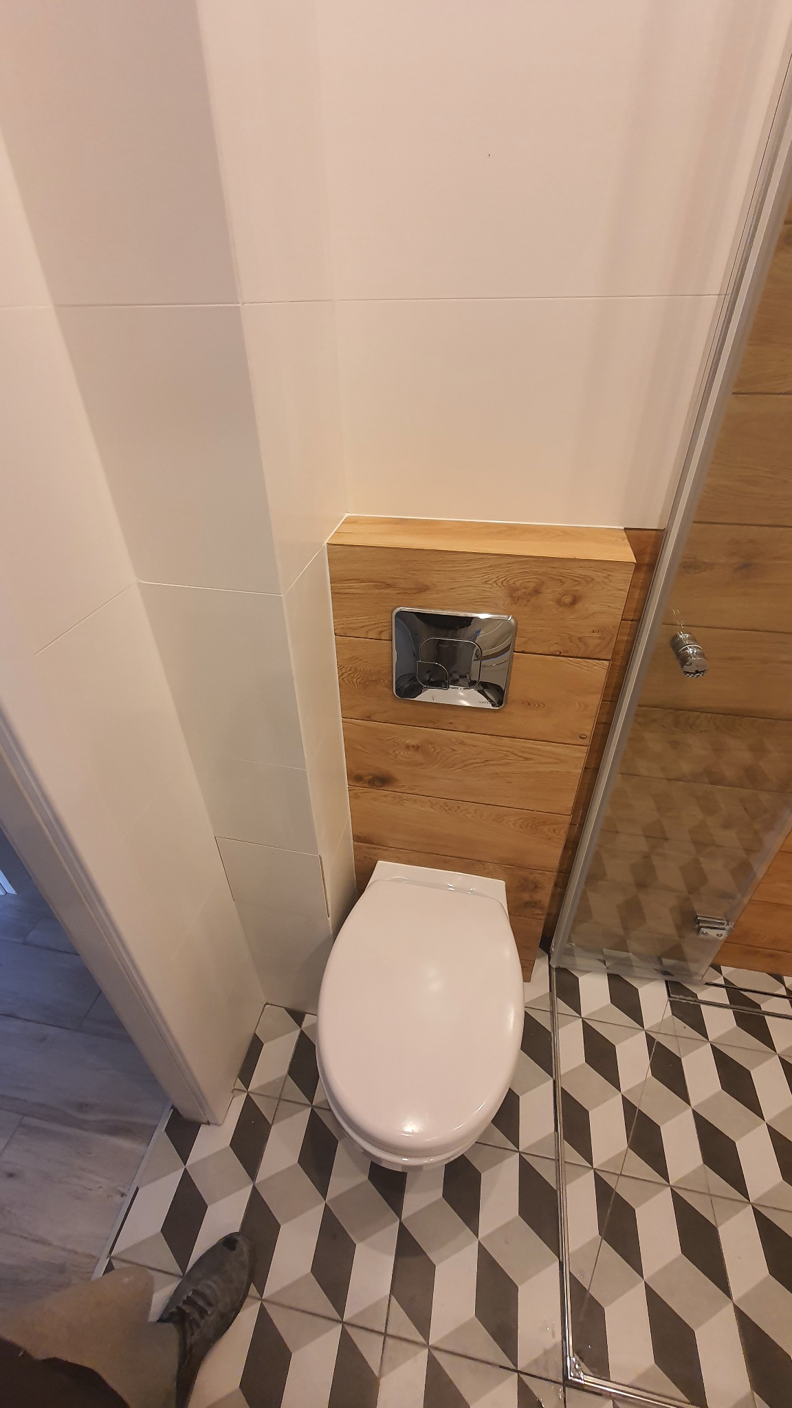 Nowoczesna toaleta podwieszana z chromowanym przyciskiem spłukującym na drewnianej obudowie, kabina prysznicowa z drewnianymi panelami i geometryczne płytki podłogowe w łazience w Krakowie, widok...