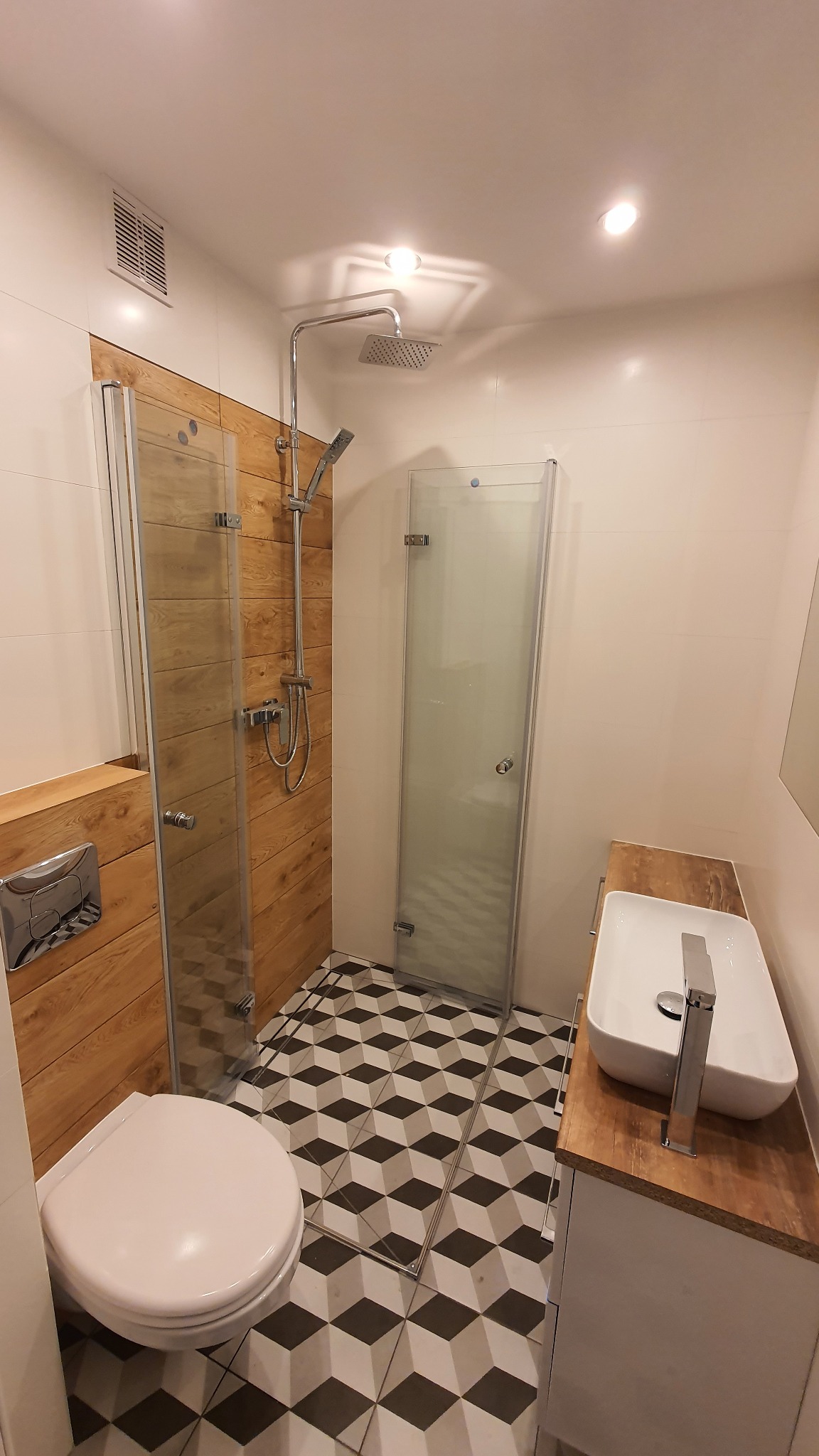 Nowoczesna łazienka po remoncie z kabiną prysznicową ze szklanymi drzwiami, umywalką nablatową na drewnianym blacie, toaletą i podłogą w geometryczny wzór 3D.