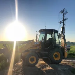 Żółto-czarna koparko-ładowarka JCB na polnej drodze, w tle wschodzące słońce i zielone pole, na ramieniu maszyny zamontowana konstrukcja przypominająca słup telekomunikacyjny.