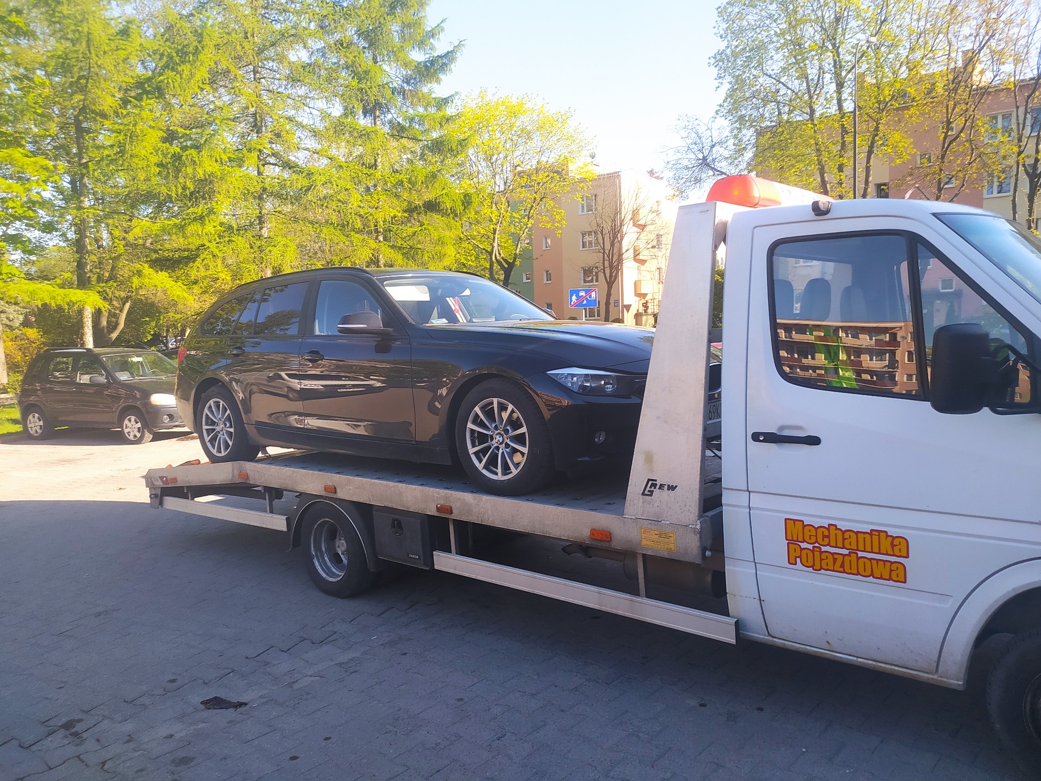 Czarny samochód kombi załadowany na platformę białej lawety z pomarańczowym światłem ostrzegawczym, zaparkowanej na brukowanym placu w słoneczny dzień, z budynkami i drzewami w tle.