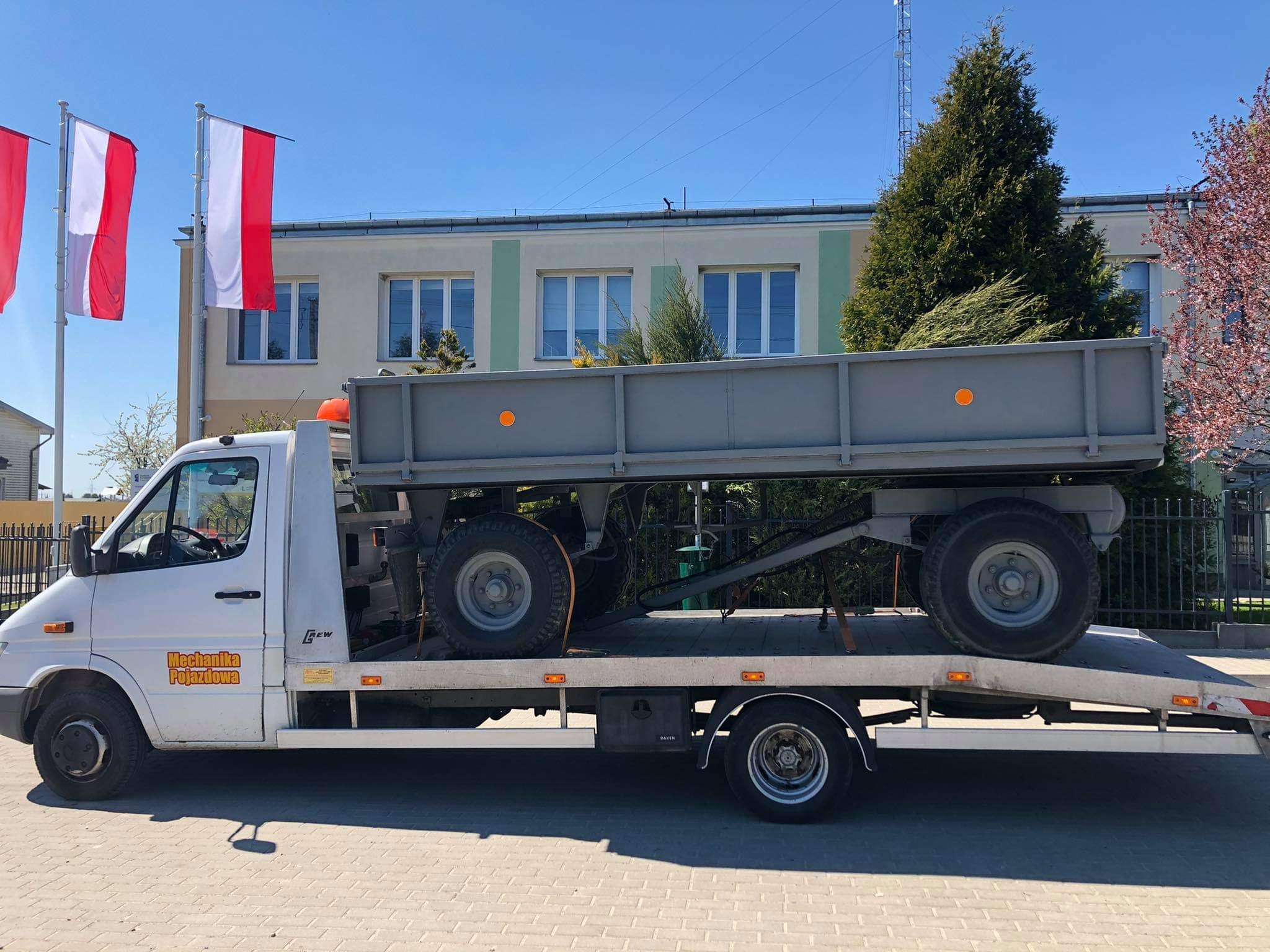 Biała laweta z przyczepą rolniczą na platformie, zaparkowana na tle budynku z polskimi flagami.
