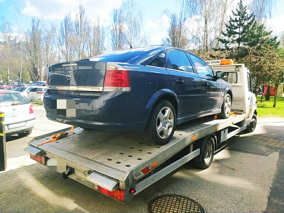 Niebieska osobówka Opel Vectra zabezpieczona pasami transportowymi na platformie lawety, widok z tyłu na parkingu z drzewami w tle.