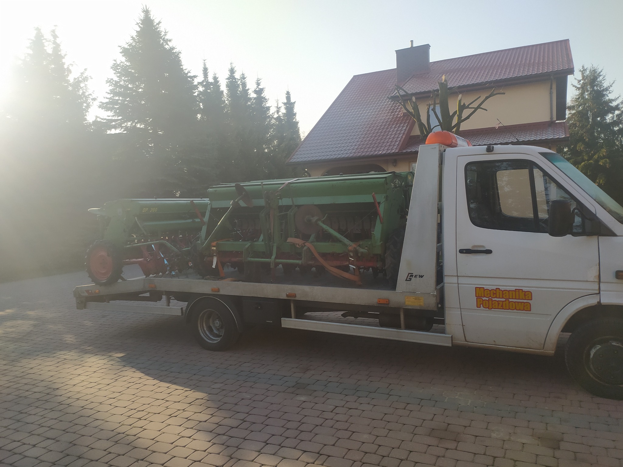 Biała laweta z pomarańczowym kogutem transportuje zielony siewnik rolniczy DP 200, zabezpieczony pomarańczowymi pasami, na tle brukowanej drogi i domu z czerwoną dachówką.