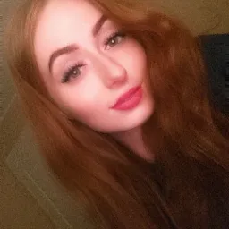 Portret kobiety z długimi, rudymi włosami i wyrazistym makijażem, skierowanej w lewo, na rozmytym tle.