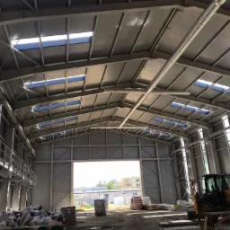 Wnętrze hali magazynowej w trakcie budowy, widoczne stalowe konstrukcje dachu z oknami dachowymi, betonowe ściany, podłoże z materiałami budowlanymi i koparka JCB w prawym dolnym rogu.