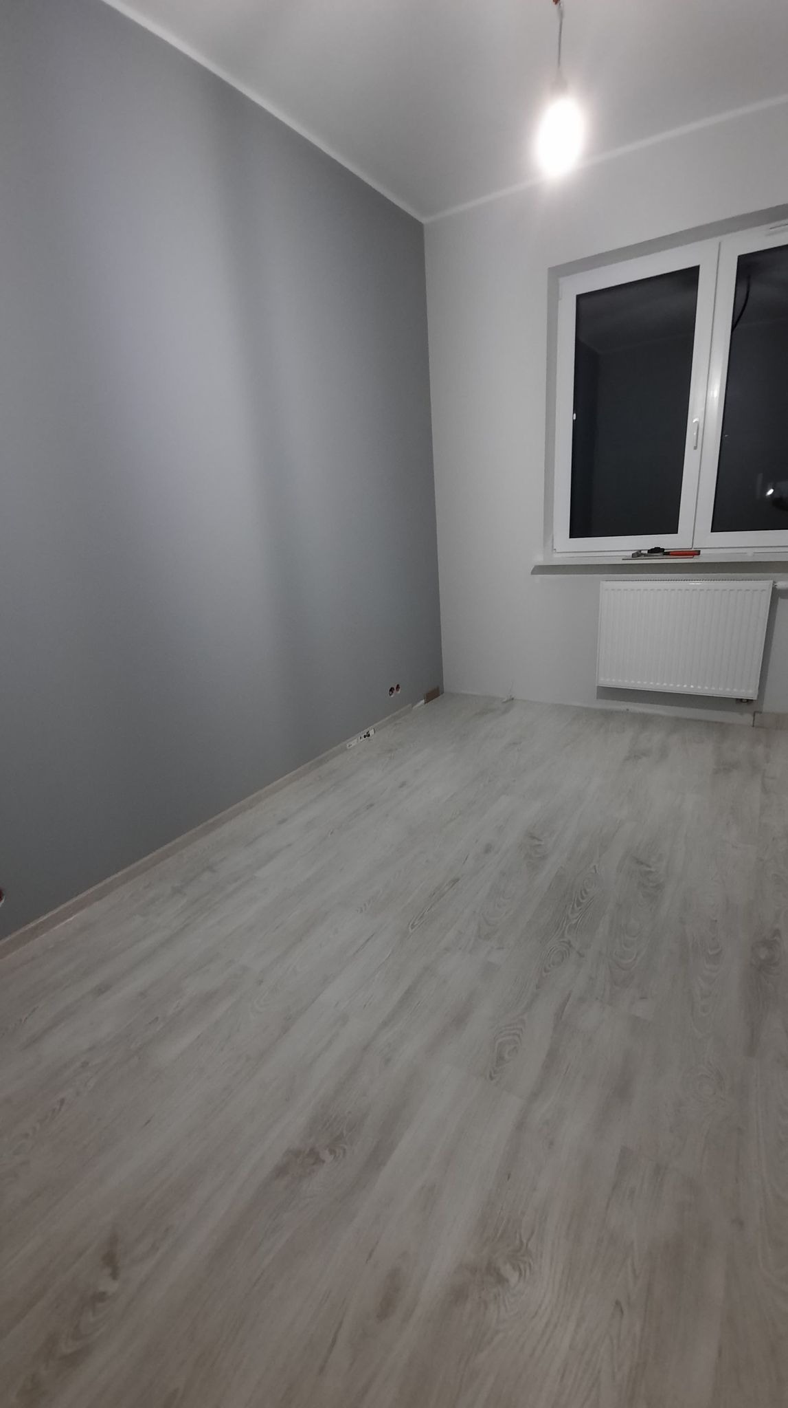 Pomalowane na szaro pomieszczenie z jasnymi panelami podłogowymi imitującymi drewno, białe okno z grzejnikiem pod spodem, widoczne puszki elektryczne w ścianie.